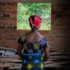 A woman in a colourful dress and headscarf looks out of a window in a brick building.
