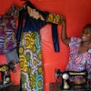 Woman smiling while seated at a sewing machine, holding up a colorful dress with yellow, blue, and green patterns and lace detailing. A second garment with purple and pink designs hangs nearby against a red fabric backdrop