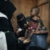  healthcare worker dressed in black clothing with a white vest is examining a small child seated on a blue plastic container. Another person wearing patterned clothing and a black face covering supports the child. The child is wearing a red and black outfit and appears thin, with visible ribs and limbs. The setting looks modest, with a brown wall in the background and another child partially visible in the foreground.
