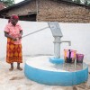 Two individuals stand at a freshly painted water pump within a walled outdoor area. One person operates the hand pump, while the other drinks from a cup. Several colourful buckets are placed on the ground near the pump, and a building made of natural materials is visible in the background.
