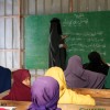 Teacher writing on chalkboard while children sit at desks in a classroom.
