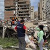 Aid worker and resident stand beside rubble as rescue teams search debris in front of a heavily damaged apartment building.