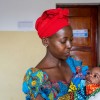 A woman wearing a bright red headscarf and colourful patterned dress cradles her baby in her arms while looking down with care. Wrapped in a vibrant blanket, the baby looks back up at her inside a healthcare facility.
