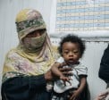 A woman wearing a patterned face covering holds a young child while standing inside a healthcare or community support centre. Another woman wearing a black face veil stands nearby, and a chart is visible on the wall in the background. The child looks toward the camera as the adults participate in what appears to be a health, nutrition, or support service visit. The image highlights maternal and child healthcare support in a community setting.