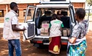 A male and female wearing concern branded jackets take boxes of soap out of the back of a car to give to a female volunteer standing beside them