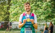 A man smiles holding his London Marathon 2025 medal after crossing the finish line. He stands proudly on grass near fencing. He is wearing a Concern branded running vest in green and pink with his name written on the front.