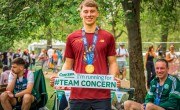 A man smiles holding a ‘I’m running for Team Concern’ sign whilst wearing his London Marathon 2025 medal after crossing the finish line. He stands proudly on grass near other runners and fencing.