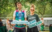 A man and woman smile together; the man is holding a ‘I’m running for Team Concern’ sign and the woman is holding a ‘#TEAM CONCERN’ sign. The man is wearing a Concern branded running vest in green and pink with his name written on the front, the woman is wearing a dark green ‘Concern Worldwide’ branded t-shirt. They stand on grass near fencing.
