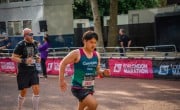 A man is looking determined as he runs towards the finish line. He is wearing a Concern branded running vest in green and pink with his name written on the front. There is another runner just behind.