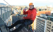 A man wearing a helmet, red jacket, and safety harness abseils down the side of a tall city building, smiling and giving a thumbs-up with one hand. The city skyline stretches into the distance beneath a bright sky.