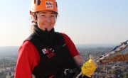 A woman is smiling as she starts her abseil off the side of a tall building. She is wearing a red T-shirt with a black body warmer and orange hard hat. She grips onto the rope with her hands in yellow gloves.