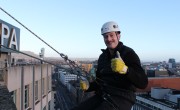 A man is smiling whilst giving a thumbs up as he starts his abseil. He is wearing a black rain jacket and white hard hat. He grips onto the rope with his hands in yellow gloves.