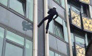 A person abseils down the side of a tall building with large windows. They are facing the building and wearing a white hard hat and navy jumpsuit.