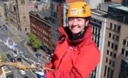 A woman is smiling as she starts her abseil off the side of a tall building. She is wearing a red rain jacket and yellow hard hat. You can see the street and cars below.