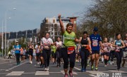 A woman smiles and celebrates, giving the camera thumbs up. She is wearing a green running t-shirt and black leggings. There are a number of runners surrounding her / close behind.  