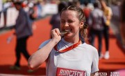 A woman smiles, biting her medal after completing the Prague Half Marathon. She is wearing a white t-shirt with her running number on the front.