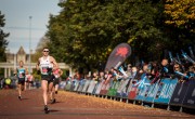 A man is looking determined as her runs on the course. He is wearing a white running vest, black shorts and sunglasses. There are spectators to the right of the runner behind a barrier, and other runners close behind