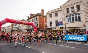 Hundreds of runners cross the start line for the Cardiff Half Marathon. All runners are wearing different coloured running vests including white, red and yellow. The start line arch is red, and spectators look on from behind a barrier.
