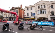 Six wheelchair users cross the start line for the Cardiff Half Marathon. All participants are wearing different colours t-shirts including navy and black, and different coloured helmets including yellow and blue. There are spectators to the right of the participants behind a barrier.