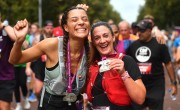  Two women smile whilst holding their Cardiff Half Marathon medal after completing the race. They are both wearing running vests, one grey and one red. 