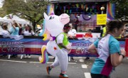 A man wearing a unicorn costume runs on the course. He is wearing a red bandana and green vest over the costume. He is running past spectators behind a barrier which reads ‘Pride Cymru’