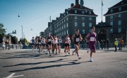 Participants for the Copenhagen Half Marathon run on the course. There are spectators behind barriers either side and a number of runners wearing different coloured running vests including black, purple and white.  