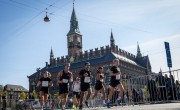 Participants for the Copenhagen Half Marathon run on the course. There are spectators behind barriers either side and a number of runners wearing different coloured running vests including black and white. They run in front of a grand building. 