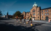 Four participants for the Copenhagen Half Marathon run on the course. They are wearing different coloured running vests including orange and yellow. They run past a grand building. 