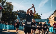  A man smiles and cheers with his hands in the air. He is wearing a grey top and black shorts with his race bib on the front. He is surrounded by other runners. They have just run under a blue arch which reads ‘Pure Gym’