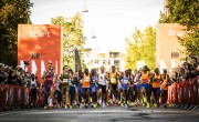 Hundreds of runners cross the start line for the Copenhagen Half Marathon. The start line branding is orange and the runners have different coloured running vests including blue, orange and white. There are a number of pacers amongst the participants. 