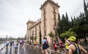 Multiple participants run on tarmac on the course. All runners are wearing different coloured running t-shirts and vests including green, red, black and blue. They are running by a large grand building which has many windows.