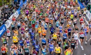 Hundreds of determined runners race on the course. There are spectators either side behind barriers, some holding black umbrellas due to the rain. The runners are all wearing their race bib on the front of their t-shirt / vest which shows their race number and name. The runners are in a wide range of coloured running t-shirts and vests including blue, yellow, orange, pink, black and green.
