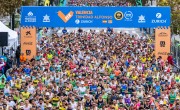 Hundreds of runners cross the start line for the Valenica Half Marathon. All runners are wearing different coloured running t-shirts and vests including green, red, black, yellow and blue. The start line arch is blue and orange with branding and advertisements across it.