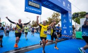 Two men in yellow and black t-shirts look ecstatic as they hold hands and cheer as they cross the finish line to celebrate completing the half marathon in 2:20:33. They are closely followed by other runners celebrating as they near the finish line. The ground is covered in blue carpet to mark the end of the race course. 