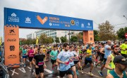 Runners cross the start line for the Valenica Half Marathon. All runners are wearing different coloured running t-shirts and vests including white, pink, black, yellow and blue. Runners are all wearing their race bib on the front of their t-shirt / vest which shows their race number and name. The weather is gloomy and the start line arch is towering above runners.