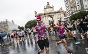 Two men pose whilst running on the course, both wearing a top saying ‘nothing can stop’ with an image of an octopus wearing sunglasses and one holding an empty plastic tumbler. It is raining, the tarmac is wet and spectators are holding umbrellas. There are more runners closely following the two men.