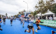 A woman with blond hair reaches her arms wide with a huge smile as she nears the finish line. She is wearing a white running vest, black shorts and has her mobile in her hand ready to take photos. She is closely followed by other runners.  