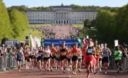 A large crowd of runners assembled at the start line of the Belfast City Marathon at Stormont Estate, with spectators lining barriers and the Stormont Parliament Buildings visible in the background under a clear blue sky.