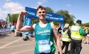 A runner wearing a Concern Worldwide vest holds a “#Team Concern” sign and a water bottle while displaying his medal after finishing the Belfast City Marathon on a sunny day.