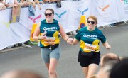 Two runners wearing green-and-yellow “Concern Worldwide” shirts run side by side in a road race, holding hands as they pass spectators behind crowd barriers. Race numbers are visible on their shirts, and banners line the street in the background.