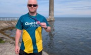 A person wearing a green-and-yellow “Concern Worldwide” running shirt stands on a rocky shoreline, holding a medal toward the camera. Calm sea water and a tall stone tower on a narrow base rise in the background under a blue sky.
