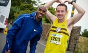 Two race participants stand on a roadside during a running event. One wears a yellow running vest with bib number 1203 and raises both arms overhead, while the other stands beside them in a blue jacket. A coach bus, stone wall, and greenery line the road in the background.
