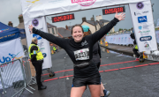 A race participant wearing black athletic clothing and bib number 7443 crosses the finish line of the Antrim Coast Half Marathon with both arms raised. A digital race clock above the finish arch shows a time of 02:58:38, with event signage, barriers, volunteers, and buildings visible along the road in the background.