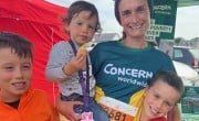 An adult wearing a green-and-yellow “Concern Worldwide” running shirt stands on grass beside a branded event tent, holding a small child on one arm, with two other children standing close. A race bib and lanyard are visible, and display boards and tents appear in the background.