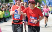 Two runners in matching red t-shirts cross the finish line together at a marathon event, with a digital timer displaying 3:42:51 overhead. Both wear race bibs and are surrounded by cheering spectators and colourful event banners under a clear blue sky.