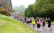 A long line of marathon runners stretches along a paved path through a scenic park on a misty morning. Participants in colourful running gear follow a route bordered by a grassy slope on the left and lush green trees on the right, with a rocky cliff face visible in the background.
