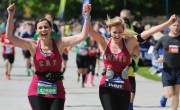 Two women runners in matching bright pink vests celebrate with arms raised and joyful expressions as they approach the finish line of a marathon. The runner on the left wears bib number 12889 with "CAT" on her vest, while the runner on the right wears bib 14681 with "ANNIE" on hers. Other participants and spectators are visible in the background under a sunny sky.