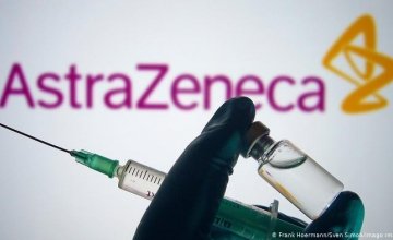 A gloved hand holds up a syringe and a vaccine vial in front of the AstraZeneca logo