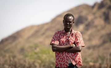 Kenyan man in red shirt standing with arms crossed against mountain background