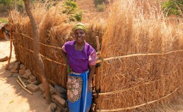Woman in purple top, blue skirt and green head wrap standing against bamboo and rushes walls 